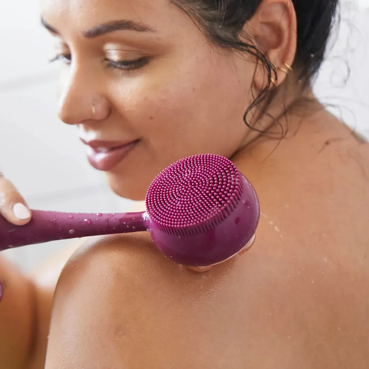 PMD Corps Propre – prix : femme souriante appliquant la brosse en silicone bordeaux sur son épaule mouillée