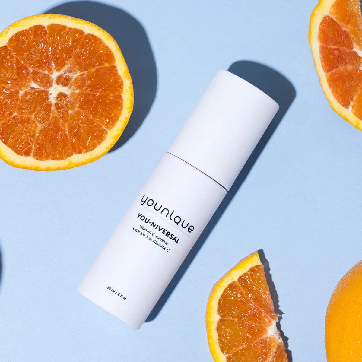 Essence à la vitamine C YOUNIQUE YOU·NIVERSAL – conseil : flacon blanc couché sur un fond bleu pervenche entouré d’oranges coupées