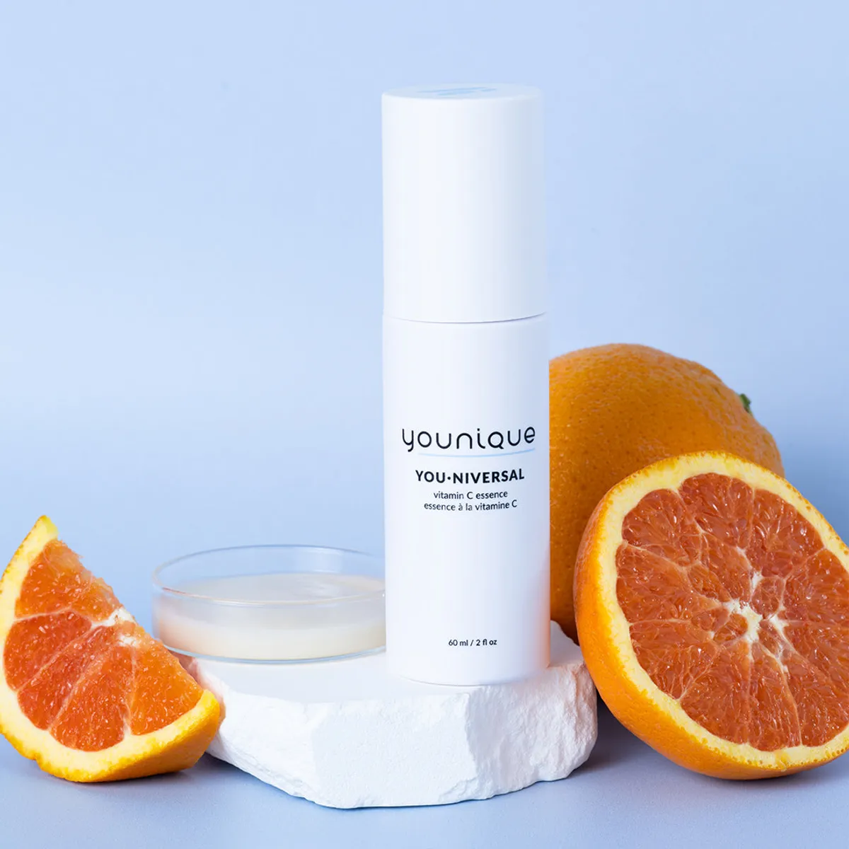 Essence à la vitamine C YOUNIQUE YOU·NIVERSAL – prix : flacon posé sur un socle en pierre claire avec des oranges et une coupelle de liquide crémeux, fond bleu clair