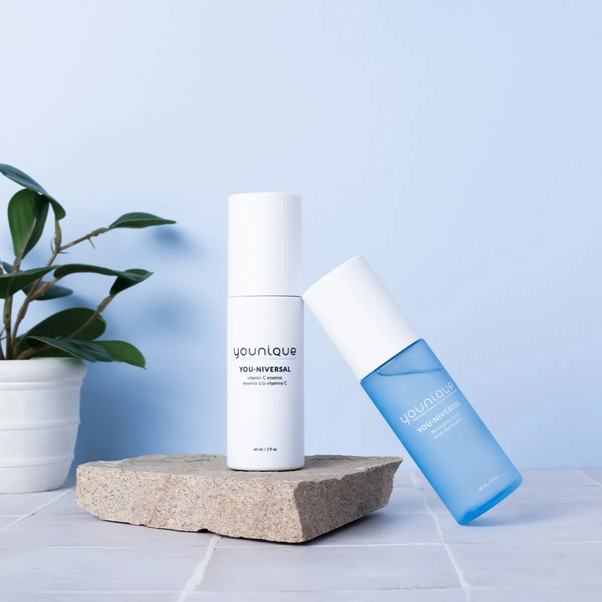 Essence à la vitamine C YOUNIQUE YOU·NIVERSAL – où acheter : flacon blanc sur une dalle de pierre à côté du flacon bleu du toner illuminateur et d’une plante verte