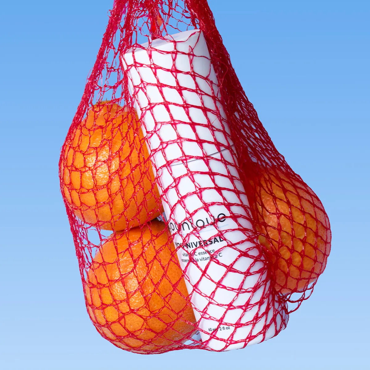 Essence à la vitamine C YOUNIQUE YOU·NIVERSAL : flacon blanc incliné dans un filet rouge avec des oranges sur fond bleu ciel, clin d’œil à la vitamine C