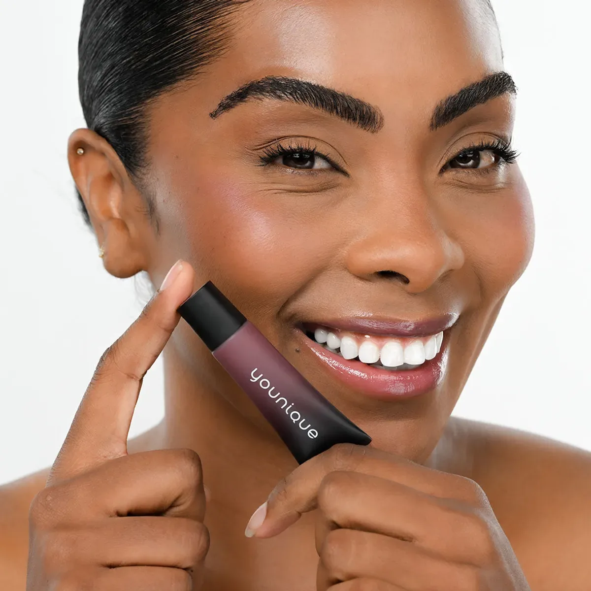 YOUNIQUE BARE·YOU lip balm : smiling model with deep skin holding the tube against her cheek, glossy mauve-brown lips