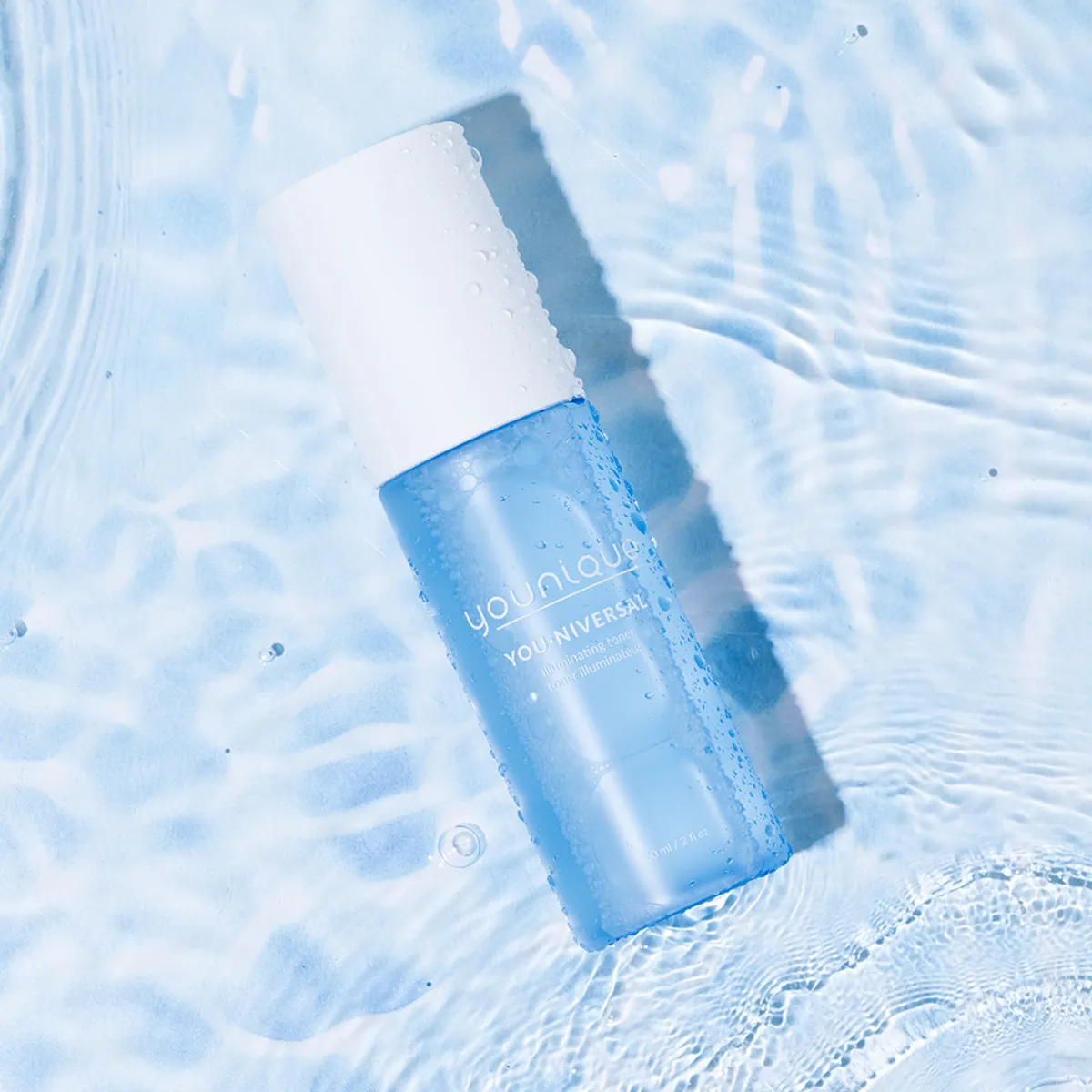 Toner illuminateur YOUNIQUE YOU·NIVERSAL – conseil : flacon couché sur une eau claire ondulée, vue du dessus avec gouttelettes