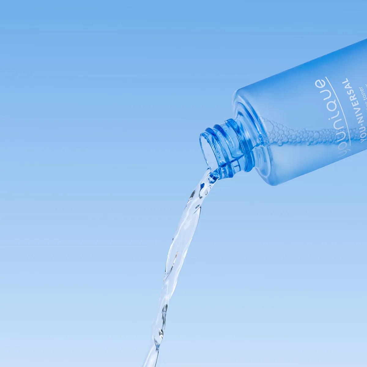 Toner illuminateur YOUNIQUE YOU·NIVERSAL : flacon ouvert versant un filet de liquide transparent sur un fond dégradé bleu ciel