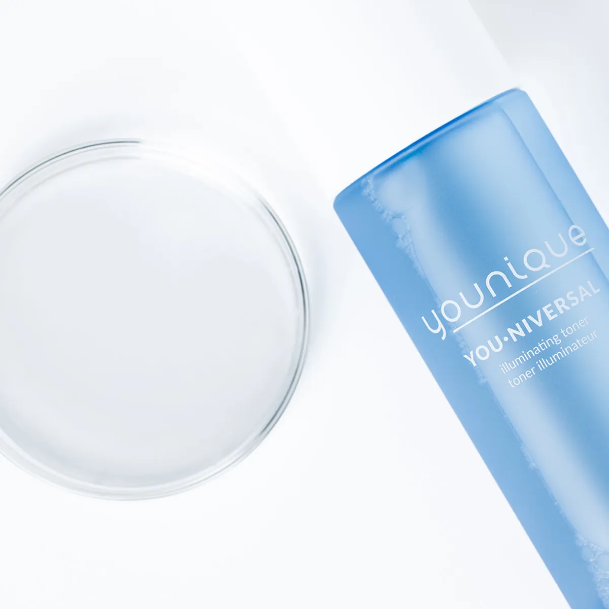 Toner illuminateur YOUNIQUE YOU·NIVERSAL : gros plan du flacon bleu givré avec une coupelle en verre remplie de toner transparent