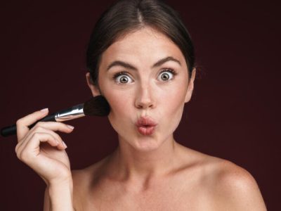 Imagen de mujer excitada semidesnuda haciendo besar los labios y usando pincel de maquillaje aislada sobre fondo rojo oscuro